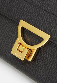 Borsa a tracolla in pelle nera con texture che presenta una chiusura geometrica in oro con ritaglio circolare e logo inciso, che mette in evidenza materiali e design.