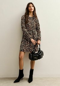 Leopard-print lange mouwen jurk met een getailleerde taille, gecombineerd met zwarte laarzen tot de knie met hakken en een zwart gewatteerde handtas.