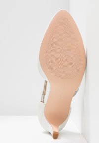 Vue de la semelle d'une chaussure blanche à talons hauts avec un bout pointu et une semelle inférieure texturée marron clair appuyée contre un mur blanc.