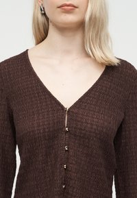 Γυναίκα που φοράει καφέ τοπ με υφή και V-λαιμόκοψη, με μικρά χρυσά κουμπιά και ασημένια σκουλαρίκια κρίκους, εμφανίζεται από τους ώμους μέχρι τη μέση.