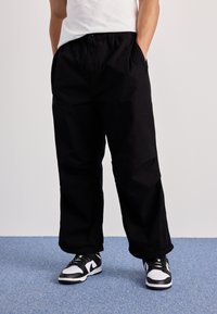 Schwarze, weit geschnittene Hosen mit elastischem Bund und seitlichen Taschen. Kombiniert mit schwarz-weißen Sneakers. Glatte Textur, lässiges Design.