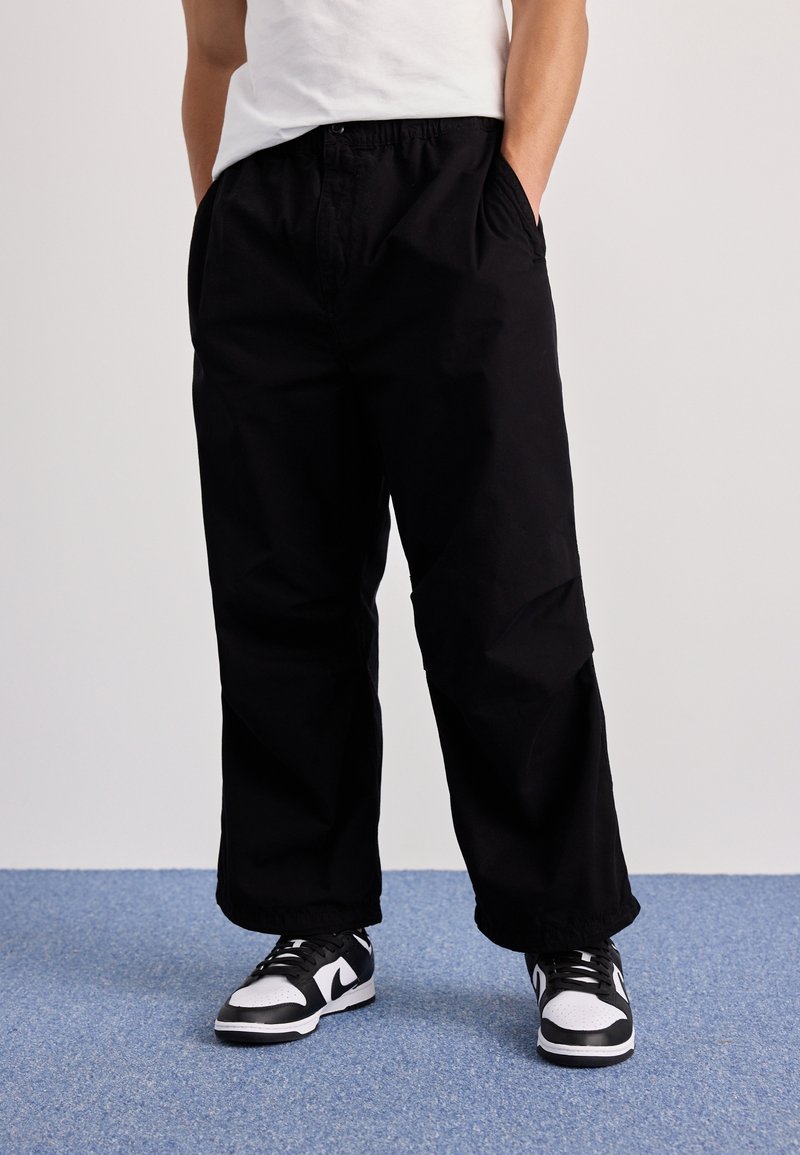 Schwarze, weit geschnittene Hosen mit elastischem Bund und seitlichen Taschen. Kombiniert mit schwarz-weißen Sneakers. Glatte Textur, lässiges Design.