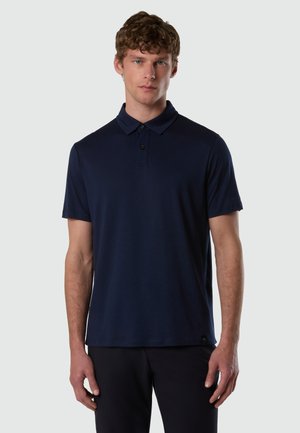 Giovane uomo con pelle chiara e capelli ricci che indossa una polo a maniche corte blu navy e pantaloni neri, in piedi contro uno sfondo semplice.