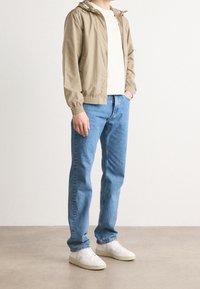 Béžová lehká bunda s kapucí, doplněná o bílé tričko a modré džíny. Bílé tenisky doplňují vzhled. Jednoduché, ležérní oblečení.