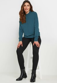 Maglione lavorato a maglia teal con colletto alto, abbinato a pantaloni skinny neri e stivaletti neri. Design semplice con polsini e orlo a costine.