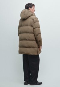 Piumino marrone con cappuccio, caratterizzato da un design trapuntato, lunghezza fino al ginocchio e una vestibilità comoda. Indossato sopra pantaloni scuri e scarpe.