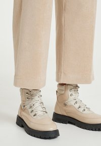 Bežové turistické boty s hladkým koženým svrškem, stříbrnými detaily a černou gumovou podrážkou. V kombinaci se světle ribbed kordovým kalhotami.