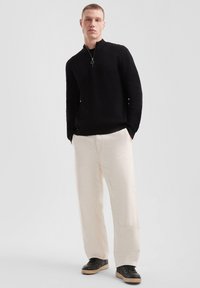 Un pull zippé noir, un pantalon large crème et des baskets noires avec une semelle beige. La tenue présente un design décontracté et des textures douces.
