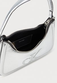 Geantă de umăr metalică argintie cu închidere cu fermoar, logo-ul „CK” embosat și căptușeală din material textil negru. Dispune de un singur mâner și cusături în contrast.