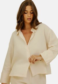 Chemise tissée beige clair avec un devant boutonné, un col classique et des manches longues. La texture semble douce et légèrement froissée.