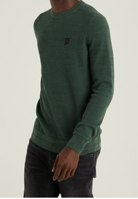 Dunkelgrüner strukturierter Pullover mit langen Ärmeln, Rundhals-Ausschnitt und einem kleinen quadratischen Logo-Patch auf der Brust, kombiniert mit dunklen Jeans.