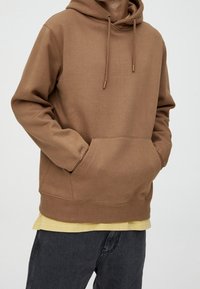 Sudadera marrón con capucha con cordón, bolsillo estilo canguro y puños acanalados. Se lleva sobre una camisa amarilla y se combina con unos vaqueros oscuros.