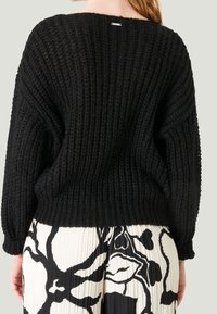 Pull en maille noir avec un motif texturé, des manches larges et une coupe décontractée. Présente une petite étiquette logo à l'arrière du cou.