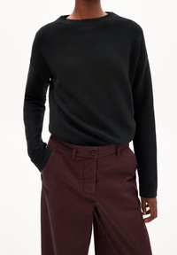 Pull en tricot noir avec un col bateau et des manches longues, associé à un pantalon bordeaux doté d'une fermeture à boutons et de poches latérales.