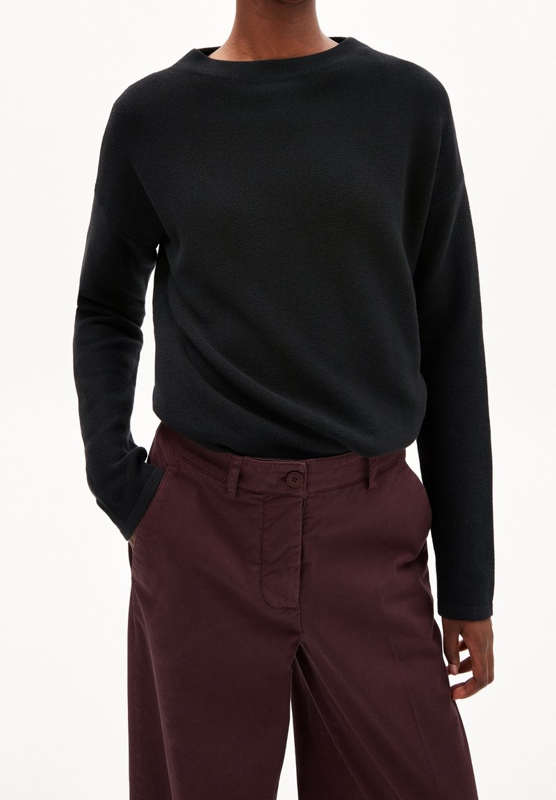 Pull en tricot noir avec un col bateau et des manches longues, associé à un pantalon bordeaux doté d'une fermeture à boutons et de poches latérales.