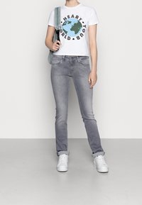Camiseta blanca con un gráfico de un globo azul y verde, combinada con jeans gris claro. Zapatillas blancas y una bolsa azul claro colgada de un hombro.
