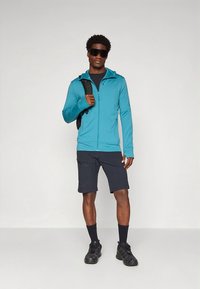 Zaļa ar rāvējslēdzēju jaka, melna T-krekls, tumši šorti, melnas zeķes un sporta apavi. Modelis valkā saulesbrilles; rokā viņam ir mugursoma.