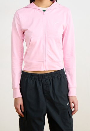 Personne portant un sweat à capuche rose clair zippé et un pantalon Nike noir avec une taille élastique, debout devant un fond clair uni.
