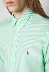 Grön- och vitrandig skjorta med knäppningkrage, med en liten lila polo-logotyp på vänster bröst. Slätt bomullstyg.