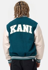 Sukienka zawodnika w kolorze teal z kremowymi skórzanymi rękawami, prążkowanym dołem i mankietami oraz wyraźnym białym napisem "KANI" na plecach.