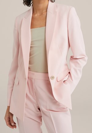 Vrouw die een lichtroze getailleerde blazer en bijpassende broek draagt, met daaronder een beige geribd mouwloos topje, hand in de zak.