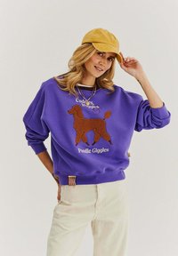 Fialová mikina s výšivkou hnědého pudla a bílým textem "Objímání, vrtění, pudlové chichotání." V kombinaci se žlutou čepicí.