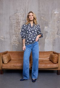 Blouse bleu marine à motifs floraux avec manches bouffantes, associée à un jean large bleu clair et des ballerines bleu foncé ; le tout devant un mur en béton.