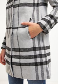 Main dans la poche du manteau, portant un manteau à carreaux gris et noir avec des boutons, associé à un jean bleu.