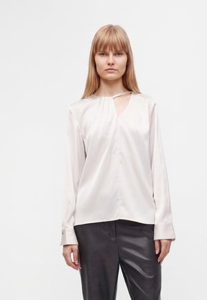 Femme aux cheveux blonds et raides porte un chemisier blanc en satin à manches longues avec une petite ouverture près de l'encolure et un pantalon en cuir noir.