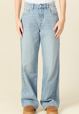 Personne portant un jean taille haute bleu clair à coupe décontractée et des chaussures en daim marron, debout devant un fond beige uni.