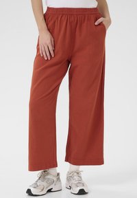 Pantalons évasés en terracotta avec une taille élastique, des poches latérales et un tissu texturé ; portés avec des chaussures de sport grises et blanches.
