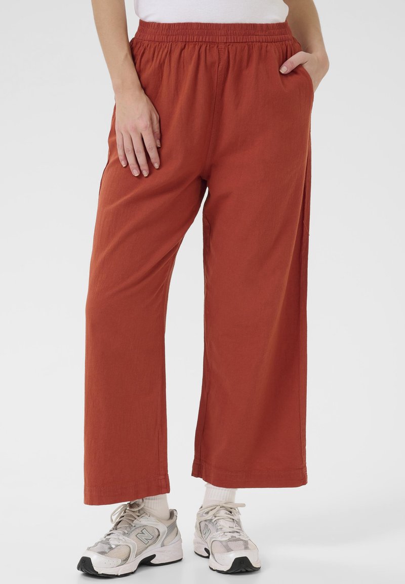 Pantalons évasés en terracotta avec une taille élastique, des poches latérales et un tissu texturé ; portés avec des chaussures de sport grises et blanches.