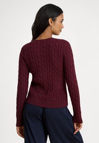 Vīnogu krāsas adīts džemperis ar pītā raksta rakstu, ribotām apmalēm un aprocēm. Aprīkots ar garām piedurknēm un apaļu izgriezumu, radīts piegulošam siluetam.