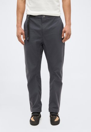 Homme portant un pantalon gris foncé avec un bouton et une boucle de ceinture noire, debout les mains le long du corps sur un fond uni.