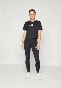 Camiseta negra corta con logo blanco y leggins negros. El modelo lleva zapatillas blancas. Textura suave, diseño atlético, forma ajustada.