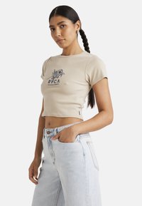 Beige cropped t-shirt in ribbed fabric with short sleeves, featuring blue floral graphic and logo on the chest. Paired with light denim jeans.