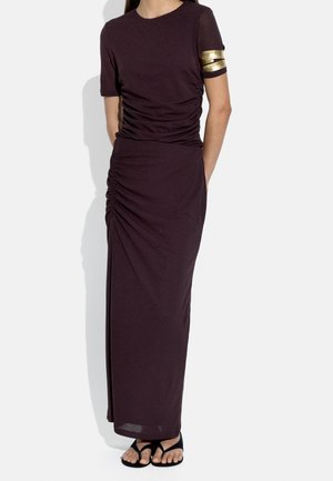 Kobieta w ciemnofioletowej sukience marszczonej z krótkimi rękawami i złotymi opaskami na górnej części ramienia, połączona z czarnymi sandałami z odkrytymi palcami.