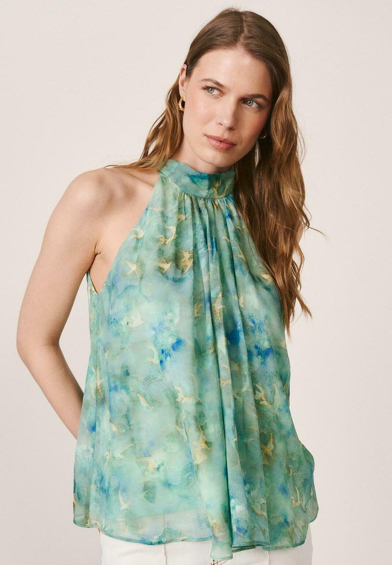 Mujer con cabello largo y castaño vistiendo un top estilo halter sin mangas con estampado floral verde azulado sobre un fondo liso.