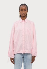 Camisa oversized listrada em rosa e branco, com colarinho, bainha elástica e mangas longas. Apresenta um bolso no peito com um detalhe de marca discreto.
