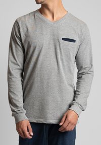 Grijze lange mouwen V-hals shirt met een gestructureerde uitstraling en een donkerblauwe patchzak op de linkerborst. Zachte stof met aansluitende manchetten.