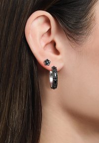 Schwarze und silberne Ohrringe mit einem großen Ring, der eingearbeitete Designs aufweist, und einem kleinen Steckerohrring mit floralen Akzenten, vor einer Hautkulisse.