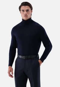 Marineblauer Rollkragenpullover aus einem weichen Stoff, schmal geschnitten, mit langen Ärmeln, kombiniert mit einem schwarzen Gürtel über maßgeschneiderten Hosen, der eine schlanke Silhouette schafft.