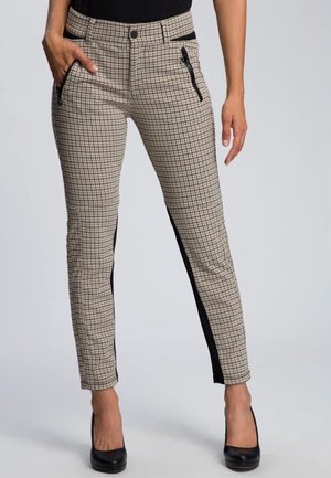 Frau trägt beigefarbene und schwarze karierte Hosen mit schwarzen Seitenstreifen, Reißverschlusstasche und schwarzen High Heels, steht vor einem grauen Hintergrund.