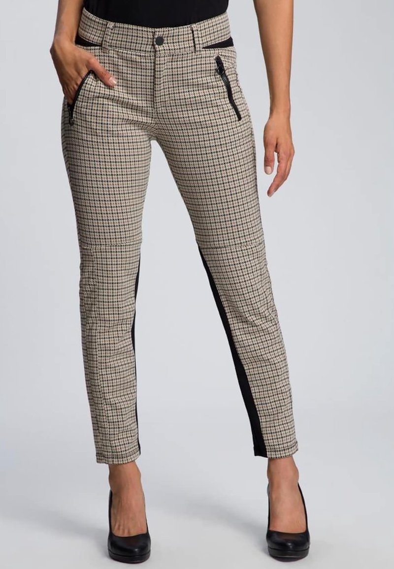 Femme portant un pantalon à carreaux beige et noir avec des panneaux latéraux noirs, une poche zippée et des talons hauts noirs, se tenant devant un fond gris.