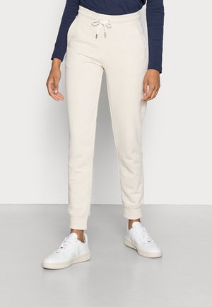 Personne portant un pantalon de jogging crème avec une taille à cordon et des baskets blanches, debout devant un fond clair uni.