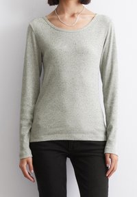 Maglietta a maniche lunghe verde chiaro con texture a costine. Presenta una scollatura ampia e sottili macchie su tutto il tessuto. Abbinata a pantaloni neri.