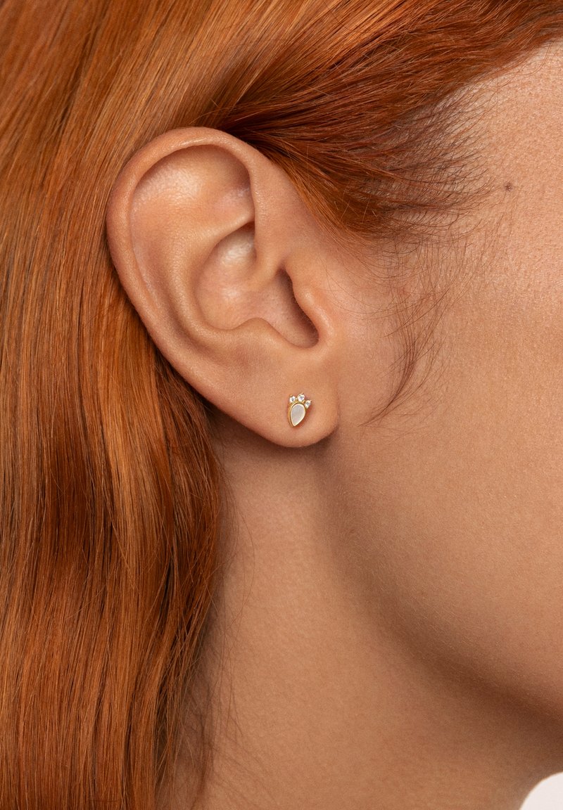 Goldfarbene Steckerohrringe mit tränenförmigem Design und drei kleinen Kristallen darüber. Positioniert am Ohr einer Person mit roten Haaren.