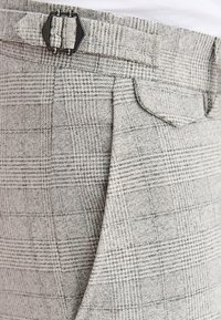 Szare spodnie w kratę z teksturowanej tkaniny, z czarną metalową klamrą na pasku i detalem tylnej kieszeni.