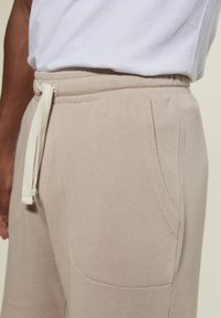 Beige Sweatpants mit Kordelzugbund und Seitentaschen, getragen mit einem weißen T-Shirt, das auf Taillen- und Oberschenkelhöhe zugeschnitten ist.