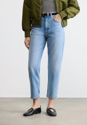 Personne portant un jean taille haute bleu clair, ceinture noire, chemise rayée, veste verte et chaussures noires pointues avec des lanières à la cheville, debout sur un sol gris.